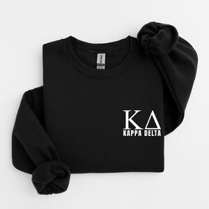Puede incluir: Una sudadera negra de cuello redondo con las letras griegas blancas KA y el texto KAPPA DELTA impresos en la parte inferior derecha del pecho. La prenda tiene mangas largas con puños acanalados y un cuello acanalado, con una etiqueta visible.