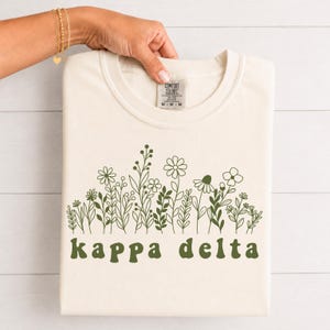 Puede incluir: Una camiseta de manga corta color crema con una ilustración de líneas en verde oscuro de varias flores silvestres en el pecho. Debajo del diseño floral, el texto kappa delta está impreso en una fuente redondeada de color verde oscuro.
