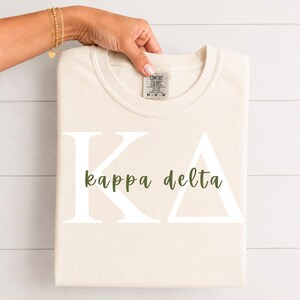Peut inclure: Un t-shirt couleur crème arborant les grandes lettres grecques blanches K et Delta, avec les mots kappa delta inscrits en écriture cursive vert foncé au centre. Le t-shirt présente une étiquette de marque Comfort Colors à l'encolure.