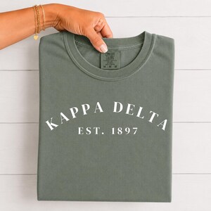 Op de afbeelding: Een olijfgroen t-shirt met ronde hals en de witte tekst Kappa Delta in een boog boven de tekst EST. 1897. Het t-shirt heeft een Comfort Colors merklabel aan de binnenkant van de kraag, dat de maat medium en 100% katoen aangeeft.