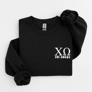 Op de afbeelding: Zwart sweatshirt met ronde hals met witte Griekse letters en de woorden "chi omega" op de voorkant. Het sweatshirt heeft lange mouwen en een geribbelde halslijn. De stof lijkt een zacht, comfortabel materiaal te zijn.