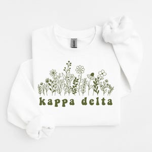 Op de afbeelding: Een wit sweatshirt met ronde hals en een donker olijfgroene lijntekening van diverse wilde bloemen op de borst. Onder het bloemmotief staat de tekst "kappa delta" gedrukt in een donker olijfgroene, afgeronde en vetgedrukte lettertype.