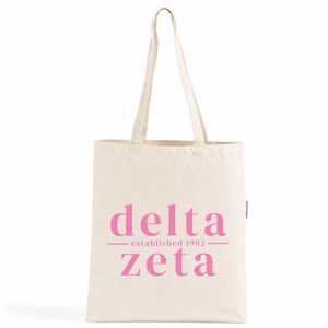 Op de afbeelding: Een natuurkleurige canvas draagtas met twee lange schouderbanden. De voorzijde van de tas is voorzien van de roze tekst "delta zeta" in een schreeflettertype, met de woorden "established 1902" gecentreerd tussen twee horizontale roze lijnen.