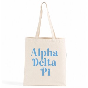 Puede incluir: Una bolsa de tela de color natural con dos asas largas para el hombro. La parte frontal de la bolsa presenta el texto Alpha Delta Pi impreso en una fuente serif de color azul claro. La bolsa tiene una forma rectangular y un diseño funcional.