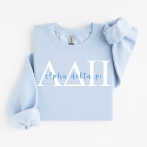 May include: A light blue sweatshirt with the Greek letters Alpha Delta Pi in white, with the words "alpha delta pi" in a lighter blue script font. The sweatshirt has a crew neck and long sleeves with rolled-up cuffs.