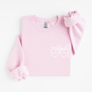 Peut inclure: Un sweat-shirt à col rond rose clair avec un motif graphique en ligne blanche sur la poitrine. Le design se compose de deux cœurs reliés attachés par un ruban, avec le texte 'kappa' dans le cœur gauche et 'delta' dans le cœur droit.