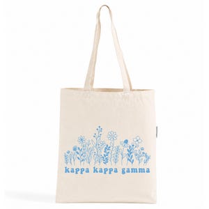 Könnte beinhalten: Eine naturfarbene Tragetasche aus Segeltuch mit zwei langen Schulterriemen. Die Vorderseite zeigt eine blaue Strichzeichnung verschiedener Wildblumen und den Schriftzug "kappa kappa gamma" in einer passenden blauen, abgerundeten Schrift.