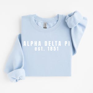 Puede incluir: Sudadera azul claro con el texto blanco "ALPHA DELTA PI est. 1851". La sudadera de cuello redondo tiene mangas largas con puños enrollados. La tela parece suave y cómoda.