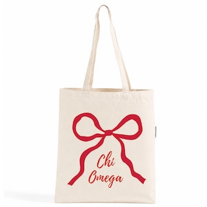 Op de afbeelding: Een natuurkleurige canvas draagtas met een grote rode afbeelding van een gestrikte strik. Onder de strik staat de tekst Chi Omega in een rood cursief lettertype. De tas heeft twee lange schouderbanden en een rechthoekig model met platte bodem.
