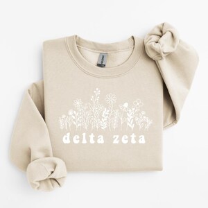 Puede incluir: Sudadera beige con diseño floral blanco y la inscripción "delta zeta". La sudadera tiene cuello redondo y mangas largas. El diseño floral está impreso en blanco.