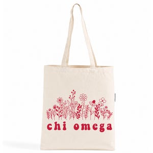 Op de afbeelding: Een crèmekleurige canvas draagtas met twee lange schouderbanden en een rode lijntekening van diverse wilde bloemen op de voorzijde. Onder het bloemendesign staat de tekst "chi omega" gedrukt in een vetgedrukt, afgerond rood lettertype.