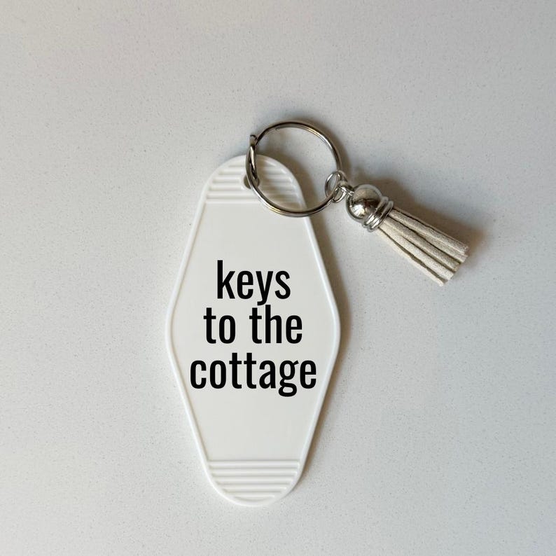 May include: A white motel-style keychain with the words "keys to the cottage" in black. The keychain has a silver metal ring and a small, light-colored tassel.