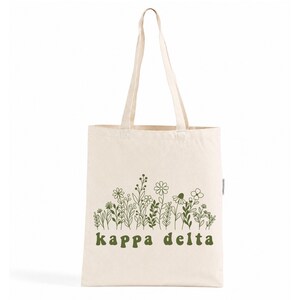Peut inclure: Un sac fourre-tout en toile couleur crème orné d'une illustration au trait vert représentant diverses fleurs sauvages et tiges. Sous le motif floral, le texte "kappa delta" est imprimé en police minuscule, grasse et arrondie, vert foncé.