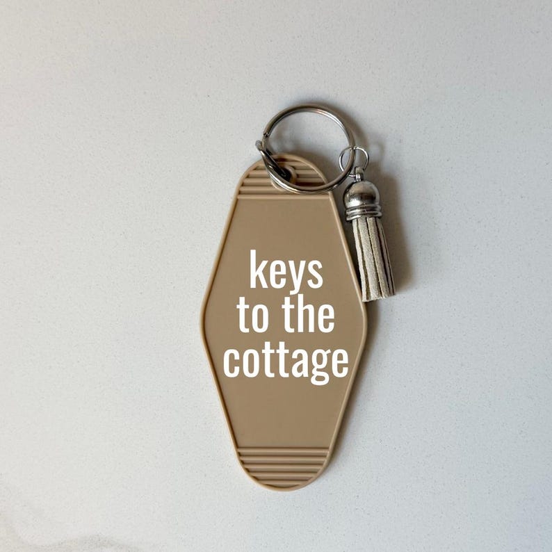 May include: A tan motel-style keychain with the words "keys to the cottage" in white. The keychain has a silver key ring and a small, off-white tassel. The background is a light, neutral color.