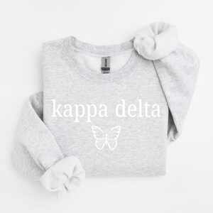 Peut inclure: Un sweat-shirt à col rond gris clair avec le texte blanc kappa delta imprimé sur la poitrine. Sous le texte se trouve un motif de papillon blanc en traits fins. Le vêtement a des manches longues avec des poignets et un col côtelés.