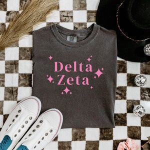 Könnte beinhalten: Ein dunkelgraues T-Shirt mit dem pinkfarbenen Schriftzug "Delta Zeta" und Sternen-Grafiken. Weiße Leinenschuhe und ein schwarzer Hut sind ebenfalls im Bild zu sehen, vor einem karierten Hintergrund.
