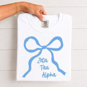 Könnte beinhalten: Ein weißes Kurzarm-T-Shirt mit einer hellblauen Grafik einer gebundenen Schleife. Unter der Schleife ist der Text Zeta Tau Alpha in einer passenden hellblauen Schreibschrift gedruckt. Das T-Shirt hat ein Comfort Colors Markenetikett am Halsausschnitt.
