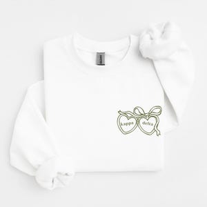 Puede incluir: Una sudadera blanca de manga larga con cuello redondo y un gráfico bordado en color verde oliva en la parte inferior derecha. El diseño muestra dos relicarios en forma de corazón unidos por un lazo, con el texto kappa y delta en su interior.