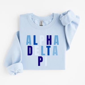 Op de afbeelding: Een lichtblauwe sweatshirt met de tekst "ALPHA DELTA PI" in blauwtinten. De sweatshirt heeft een ronde hals en lange mouwen met opgerolde manchetten. De stof ziet er zacht en comfortabel uit.