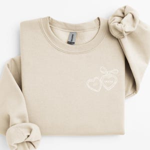Könnte beinhalten: Beiges Sweatshirt mit Rundhalsausschnitt und weißen, gestickten Herzen mit den Worten "Chi" und "Omega". Das Sweatshirt hat einen gefalteten Ärmel und ein Gildan-Etikett. Das Design ist auf der Brust zentriert.