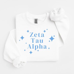 Peut inclure: Un sweat-shirt blanc à manches longues et col rond avec le texte Zeta Tau Alpha en police serif bleu clair. Le motif est entouré de diverses icônes d'étoiles à quatre branches de différentes tailles, centrées sur le devant du vêtement.