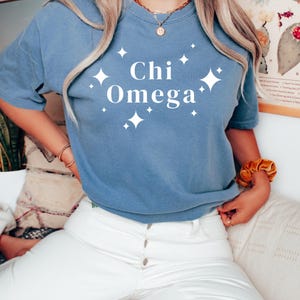 Könnte beinhalten: Ein blaues T-Shirt mit dem weißen Schriftzug "Chi Omega" und weißen Sternen. Das Shirt wird mit einer weißen Hose kombiniert. Das Bild vermittelt einen entspannten, alltäglichen Stil, der Komfort und Schlichtheit betont.