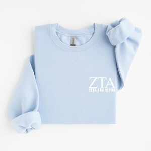 Puede incluir: Una sudadera de cuello redondo en color azul claro con el texto blanco ZTA y Zeta Tau Alpha impreso en la parte inferior derecha. La prenda tiene mangas largas con puños acanalados y cuello acanalado, con una etiqueta Gildan en el cuello.