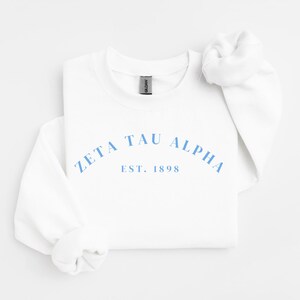 Peut inclure: Un sweat-shirt blanc à col rond avec le texte bleu clair Zeta Tau Alpha arqué au-dessus du texte Est. 1898. Le vêtement possède des manches longues avec des poignets côtelés et une étiquette de marque Gildan à l'intérieur du col.