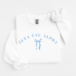 Puede incluir: Una sudadera blanca de cuello redondo con el texto Zeta Tau Alpha en una fuente arqueada de color azul claro. Debajo del texto hay un gráfico de color azul claro de un lazo. La prenda tiene mangas largas con puños acanalados y una etiqueta Gildan.