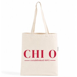 Peut inclure: Un sac fourre-tout en toile de couleur crème doté de deux longues anses. L'avant du sac présente le texte rouge CHI O centré au-dessus de la mention established 1895, encadrée par deux lignes horizontales rouges de chaque côté.