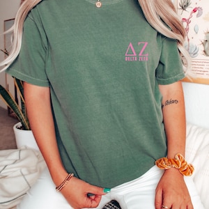 Op de afbeelding: Een olijfgroen t-shirt met de Griekse letters ΔZ en de woorden DELTA ZETA in roze gedrukt. Het shirt met korte mouwen is effen van kleur. De afbeelding toont het gedragen shirt.