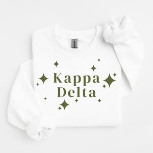 Puede incluir: Una sudadera blanca de manga larga con cuello redondo que presenta el texto Kappa Delta en verde oliva oscuro centrado en el pecho. El texto está rodeado por formas de estrellas de cuatro puntas de varios tamaños en el mismo color.