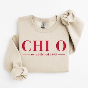 Peut inclure: Un sweat-shirt crème avec le texte rouge "CHI O" au-dessus de "established 1895". Le sweat-shirt a un col rond et des manches longues, avec une manche repliée. L'étiquette de la marque Gildan est visible.