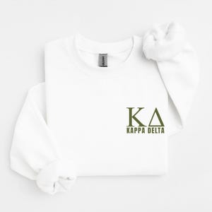 Op de afbeelding: Een wit sweatshirt met lange mouwen en een ronde hals, voorzien van de olijfgroene Griekse letters Kappa Delta en de tekst KAPPA DELTA op de rechterborst. Het kledingstuk heeft een standaard geribbelde halslijn en een Gildan-label in de kraag.