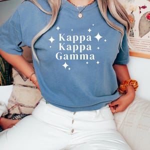 Op de afbeelding: Een blauw T-shirt met de witte tekst "Kappa Kappa Gamma" en sterren. Dit casual shirt is gemaakt van een zacht materiaal. Het shirt wordt gedragen met een witte broek.