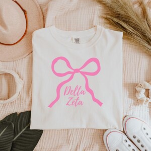 Peut inclure: T-shirt blanc avec un motif de nœud rose et l'inscription "Delta Zeta". Le t-shirt est posé à plat sur une surface texturée. Des baskets blanches sont visibles en bas à droite.
