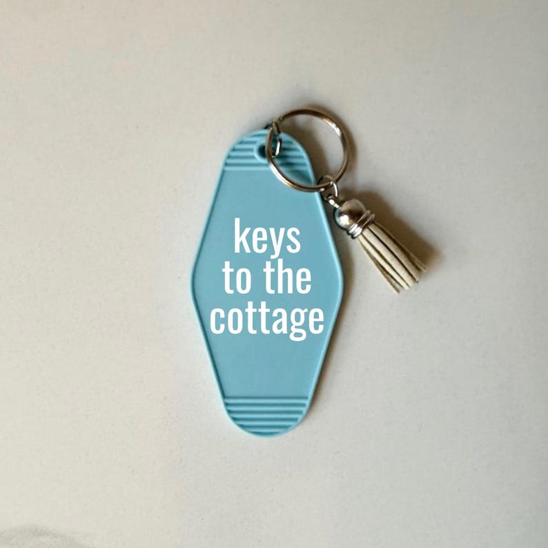 May include: A light blue motel-style keychain with the words "keys to the cottage" in white. The keychain has a silver ring and a cream-colored tassel. The background is a light, neutral color.