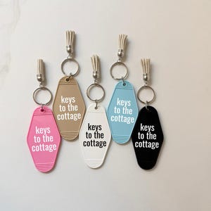 May include: Five motel-style keychains in pink, tan, white, light blue, and black. Each keychain has a silver ring and a tassel. The text "keys to the cottage" is printed on each keychain.