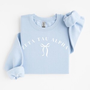 Peut inclure: Un sweat-shirt à col rond bleu clair avec le texte blanc Zeta Tau Alpha arqué au-dessus d'un graphique blanc représentant un nœud ruban. Le vêtement à manches longues possède un col et des poignets côtelés, avec une étiquette Gildan visible.