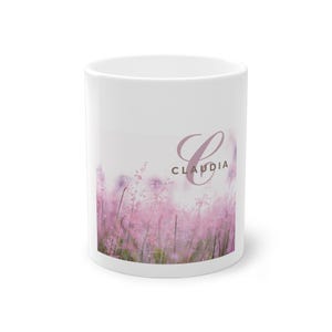 Op de afbeelding: Witte keramische mok met een bloemenontwerp van roze bloemen en de naam "Claudia". De mok heeft een gebogen handvat en een glad oppervlak. De tekst is in een gestileerd lettertype.