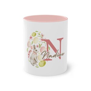 Könnte beinhalten: Weißer Keramikbecher mit rosa Innenraum. Der Becher hat ein Ostermotiv mit einem Hasen, Eiern, Blumen und dem Buchstaben "N" in Korallenfarbe. Der Name "Nadine" ist in einer Schreibschrift geschrieben.