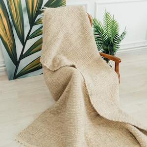 Könnte beinhalten: Eine beige gewebte Wurfadecke mit Fransen ist über einen Holzstuhl drapiert. Die Decke hat ein strukturiertes Aussehen und besteht aus einem dicken, weichen Material. Der Hintergrund umfasst ein Gemälde mit grünen und gelben Blättern.