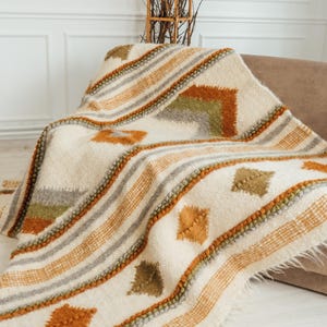 Può includere: Coperta tessuta color crema con un motivo geometrico nei toni dell'arancione, grigio e verde. La coperta ha un bordo sfrangiato ed è drappeggiata su un divano marrone. Il design presenta strisce e diamanti.