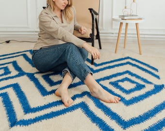May include: A cream rug featuring a geometric blue design. The rug is placed on a light floor. A person is sitting on the rug, wearing jeans and a long-sleeved top. A black lamp and a small side table are also visible in the room.