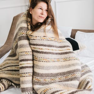 May include: A cream, brown, and olive green striped blanket. The blanket is wrapped around a person on a bed with white sheets. The blanket has a textured appearance and a fringed edge. The bed is in a room with white walls.