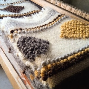 May include: A close-up of a handwoven textile on a wooden loom. The textile has a cream-coloured base with textured brown, grey, and yellow geometric patterns. The piece is made of thick, soft yarn, showcasing a rustic, handcrafted aesthetic.