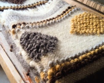 May include: A close-up of a handwoven textile on a wooden loom. The textile has a cream-coloured base with textured brown, grey, and yellow geometric patterns. The piece is made of thick, soft yarn, showcasing a rustic, handcrafted aesthetic.