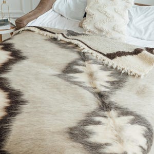 Könnte beinhalten: Eine Bettdecke in neutralen Tönen mit einem geometrischen Muster in Grau und Braun. Die Decke ist über ein Bett mit einem weißen Kissen und einem dekorativen Kissen mit Zickzackmuster drapiert. Der Raum ist hell.