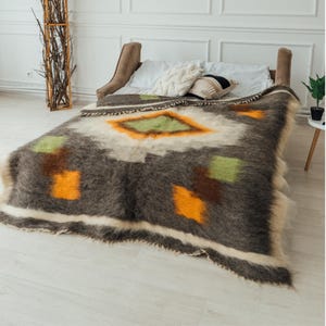 May include: A large, rectangular blanket with a geometric pattern in shades of brown, orange, green, and white. The blanket is draped over a bed, showcasing its textured, fluffy appearance. The design features diamond and square shapes.