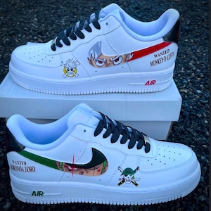 Könnte beinhalten: Weiße Turnschuhe mit schwarzen Schnürsenkeln und Anime-Grafiken. Ein Schuh hat einen roten Streifen mit dem Text "WANTED MONKEY-D-LUFFY" und eine Charakterillustration. Der andere hat einen grünen Streifen mit "WANTED RORONOA ZORO".